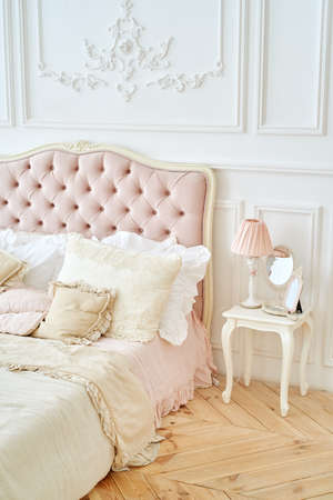 Elegant home or hotel bedroom interior. Pillows on luxury buttoned bed and  pink lamp, mirror on white wooden table, vintage styleの写真素材