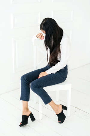 Full length portrait of young woman with long hair in white shirt sitting on chair on white background in studioの写真素材