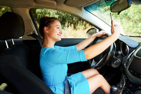 Happy young woman holding mobile phone and taking photos while driving a car. Smiling girl taking selfie picture with cellphone camera outdoors in car. Holidays and tourism conceptの写真素材