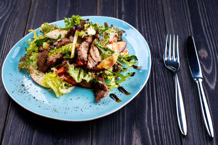 Veal salad with lettuce, apple, pear and tomato on blue plate on dark wooden background, copy space. Restaurant food, close up. Meat saladの写真素材