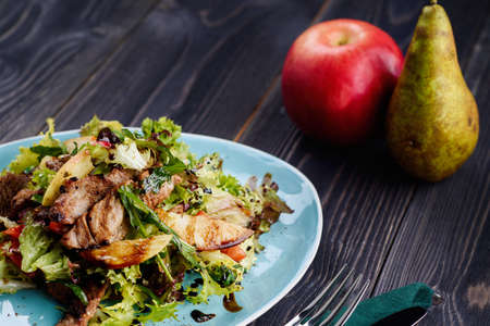Veal salad with lettuce, apple, pear and tomato on blue plate on dark wooden background, copy space. Restaurant food, close up. Meat saladの写真素材