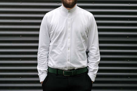 Confident young bearded man in white shirt keeping arms in pockets while standing against grey metal backgroundの写真素材