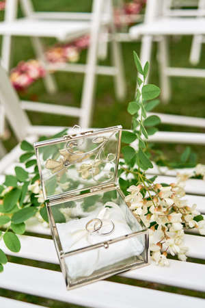 Two golden wedding rings in glass box and branch of white acacia with flowers and leaves on wooden surface.の写真素材