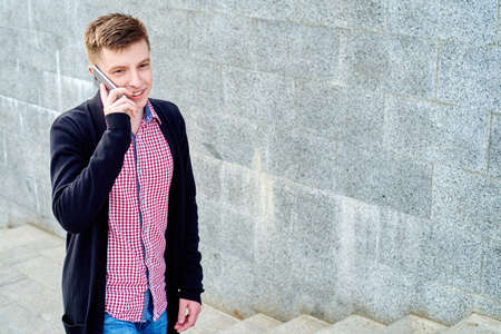 Cheerful young man in plaid shirt and jacket walking up the stairs and talking on cellphone outdoors, free space.の写真素材