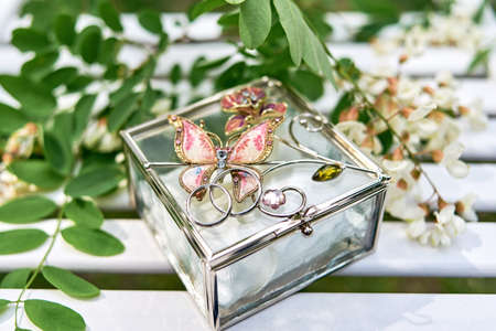 Two golden wedding rings on glass box with butterfly and branch of white acacia with flowers and leaves on wooden surface.の写真素材