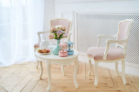 Two pink soft chairs and white wooden round table with breakfast, vase with flowers and merry-go-round musical carousel with horses.の写真素材