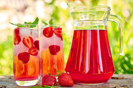 Strawberry lemonade, cocktail with ice and mint in glasses and jug with green background outdoors,...