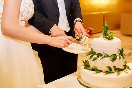 Bride and groom cutting elegant white multi level wedding cake decorated with fresh green leaves on table in restaurant with gold interior, copy spaceの写真素材