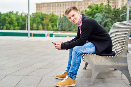 Young man with smartphone sitting on city street bench, copy space. Portrait of student using mobile phone and looking at camera outdoor, side view.の写真素材