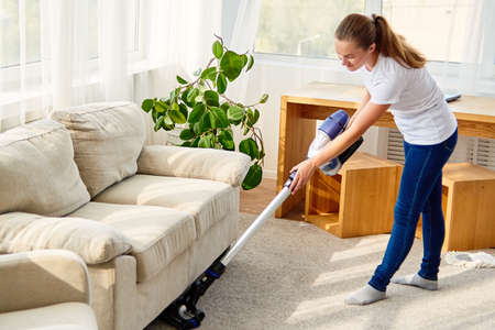 Full length body portrait of young woman in white shirt and jeans cleaning carpet with vacuum cleaner in living room, copy space. Housework, cleanig and chores conceptの写真素材