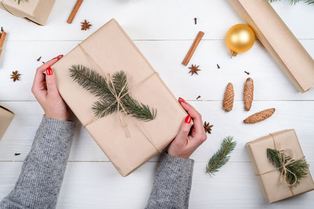 Woman with red manicure holding Christmas present wrapped in kraft paper on white wooden table background, free space. Close up of gift box in girl's hand. Christmas background. Flat lay, top viewの写真素材