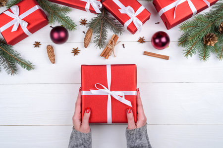 Woman holding red Christmas present laying on white wooden background. Christmas background with red gift boxes, fir tree branches, pine cones, cinnamon sticks and stars anise. Flat lay, top viewの写真素材