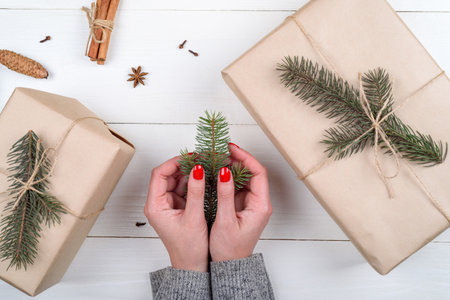 Christmas gift wrapping background, copy space. Female hands holding branch of fir tree, gift boxes wrapped in kraft, top view. Winter holidays concept, flat lay. Woman with red manicure packing giftの写真素材
