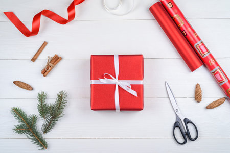 Top view of red Christmas gift box, wrapping paper, ribbon, fir tree branches, cones, cinnamon and scissors on white wooden background, copy space. Gift wrapping, packing. Flat lay, free spaceの写真素材
