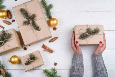 Christmas gift wrapping background, copy space. Female hands holding christmas present box wrapped in kraft, top view. Winter holidays concept, flat lay. Woman with red manicure packing Christmas giftの写真素材