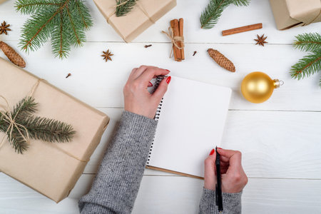 Top view of woman hands with pen writing goals or to do list in blank notebook near Christmas gift boxex on wooden background, free space. Flat lay. Holiday gift boxes wrapped in kraftの写真素材
