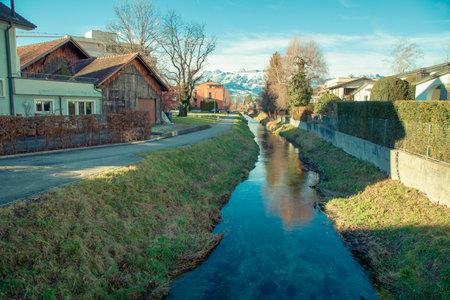 Alps mountains in Liechtenstein. Medieval Red House, calm narrow mountain river and jogging track, on the background of residential buildings, blue sky and snow-capped mountains. Liechtenstein, Vaduzの写真素材