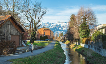 Happy girl in Alps mountains in Liechtenstein. Medieval Red House, calm narrow mountain river and jogging track, residential buildings, blue sky and snow-capped mountains. Liechtenstein, Vaduzの写真素材