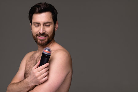 Positive and shirtless bearded man looking at deodorant isolated on gray with copy space