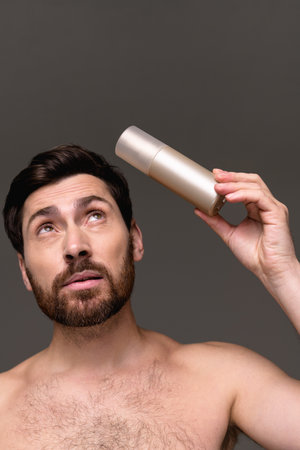 Pensive shirtless man holding hairspray isolated on gray backgroundの写真素材