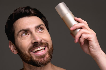 Smiling bearded man holding hairspray isolated on grey background