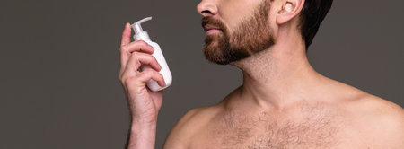 Banner crop of shirtless bearded man holding cosmetic lotion isolated on grey backgroundの写真素材