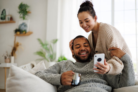 Smiling young Asian woman in jumper showing smartphone to African American boyfriend with coffee while relaxing at homeの写真素材