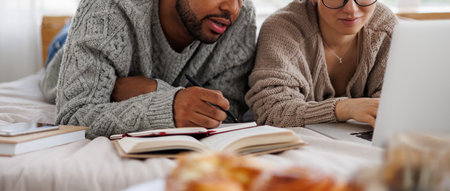 banner shot of interracial couple in sweaters using laptop and books during online education on bed...