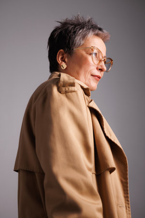 Confident mature woman in stylish eyeglasses and trench coat standing isolated on grayの写真素材