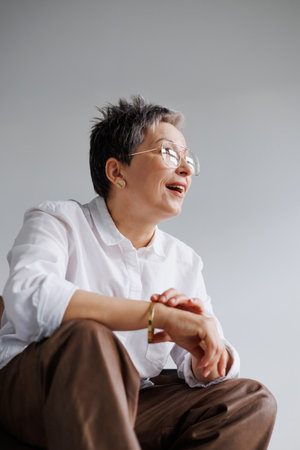 Cheerful and elegant mature woman in eyeglasses and white shirt touching golden bracelet isolated on grayの写真素材