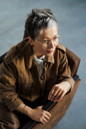 Mature woman in eyeglasses and casual jacket sitting on armchair on gray background