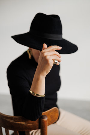 Stylish mature woman in golden accessories and black turtleneck touching hat on gray