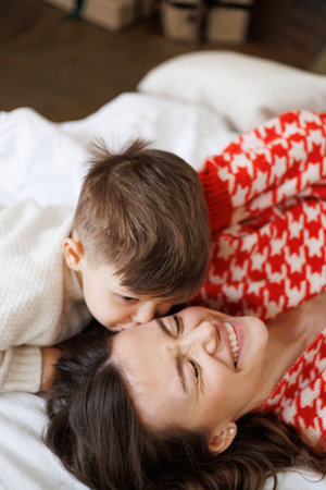Toddler boy in knitted sweater kissing cheerful mother on bed at home