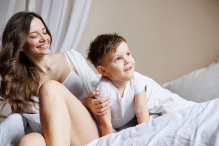 Cheerful toddler boy in pajama sitting on bed near smiling mom at homeの写真素材