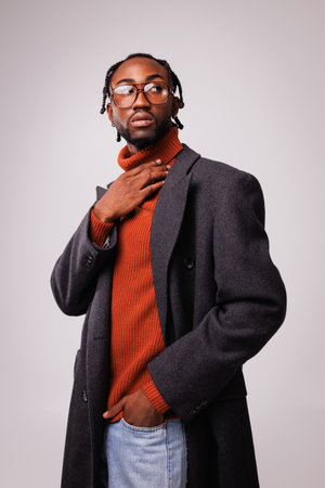 Confident and fashionable african american man in eyeglasses, classic coat and jeans touching red sweater isolated on grayの写真素材