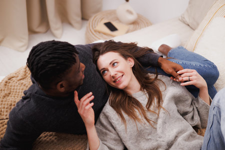 Smiling woman holding hand of African American boyfriend while relaxing on bedの写真素材