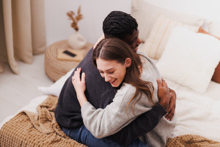 Joyful multiethnic couple in casual clothes hugging on bed at home