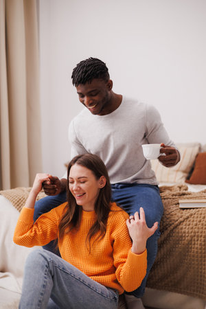 Smiling african american man holding hand of girlfriend and coffee in bedroom