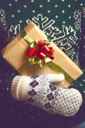 Girl holds in hands Christmas present. Christmas background. Gifts for men. Knitted mittens. Knitted dress. Box with gifts. Merry Christmas. Toned image.の写真素材
