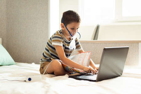 Stay at home quarantine coronavirus pandemic prevention. A child learns home lessons in an oxygen mask with a nibulizer, laptop and a book during quarantine. Prevention epidemic. COVID-19.の写真素材