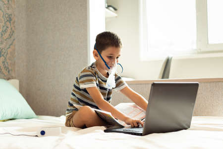 Stay at home quarantine coronavirus pandemic prevention. A child learns home lessons in an oxygen mask with a nibulizer, laptop and a book during quarantine. Prevention epidemic. COVID-19.の写真素材