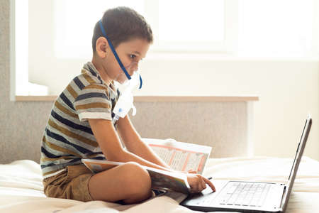 Stay at home quarantine coronavirus pandemic prevention. A child learns home lessons in an oxygen mask with a nibulizer, laptop and a book during quarantine. Prevention epidemic. COVID-19.の写真素材
