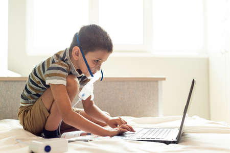 Stay at home quarantine coronavirus pandemic prevention. A child learns home lessons in an oxygen mask with a nibulizer, laptop and a book during quarantine. Prevention epidemic. COVID-19.の写真素材
