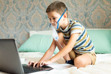 Stay at home quarantine coronavirus pandemic prevention. A child learns home lessons in an oxygen mask with a nibulizer, laptop and a book during quarantine. Prevention epidemic. COVID-19.の写真素材