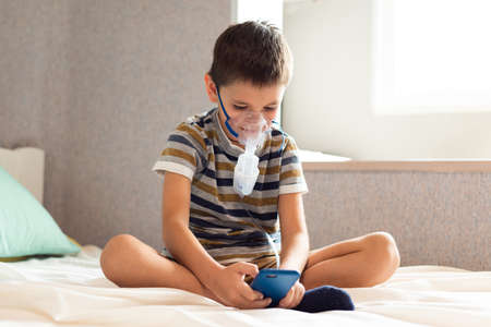 Stay at home quarantine coronavirus pandemic prevention. A child in an oxygen mask with a nibulizer plays on a mobile phone during quarantine. Prevention epidemic. COVID-19.の写真素材