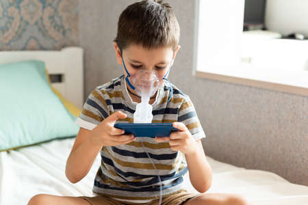 Stay at home quarantine coronavirus pandemic prevention. A child in an oxygen mask with a nibulizer plays on a mobile phone during quarantine. Prevention epidemic. COVID-19.の写真素材