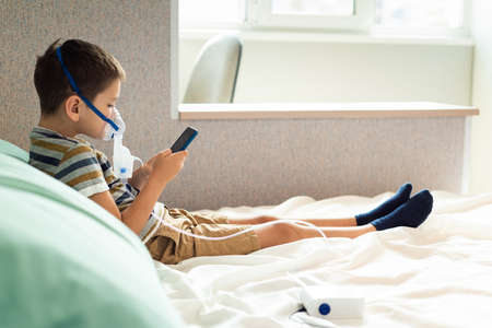 Stay at home quarantine coronavirus pandemic prevention. A child in an oxygen mask with a nibulizer plays on a mobile phone during quarantine. Prevention epidemic. COVID-19.の写真素材