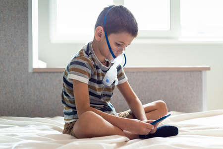 Stay at home quarantine coronavirus pandemic prevention. A child in an oxygen mask with a nibulizer plays on a mobile phone during quarantine. Prevention epidemic. COVID-19.の写真素材