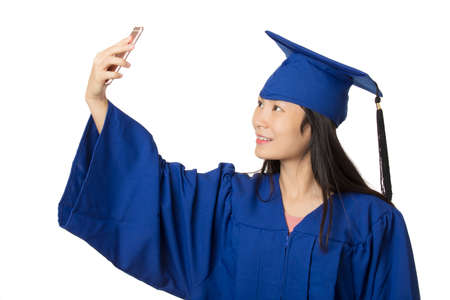 Beautiful Asian woman using a smartphone to take a selfie of herself wearing a blue graduation gown isolated on white backgroundの写真素材