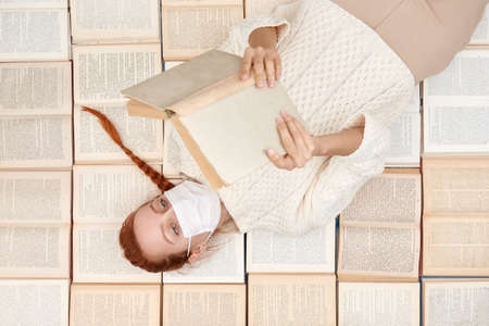 A girl is lying among books and reading a book. A student in a protective mask is reading a book at home. Closing schools and universities. Home quarantine concept.の写真素材